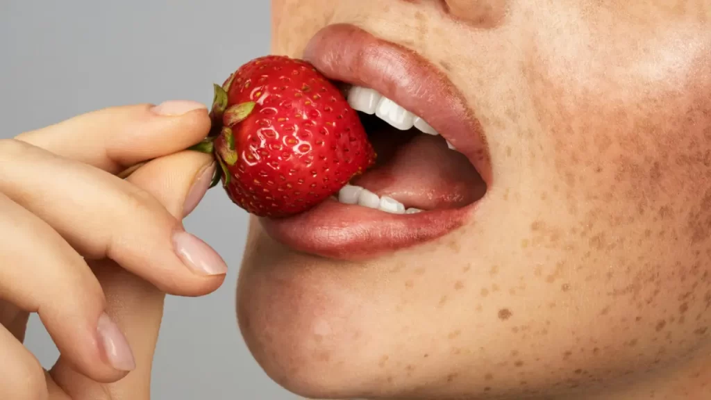 Mulher com sardas saboreando um morango, destacando os benefícios do morango para a pele.
