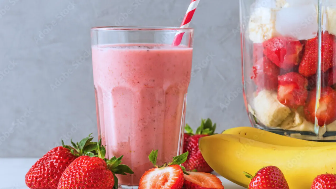 Copo de smoothie de morango com canudo listrado, rodeado por morangos frescos e bananas, ao lado de um liquidificador com pedaços de morango, banana e gelo.