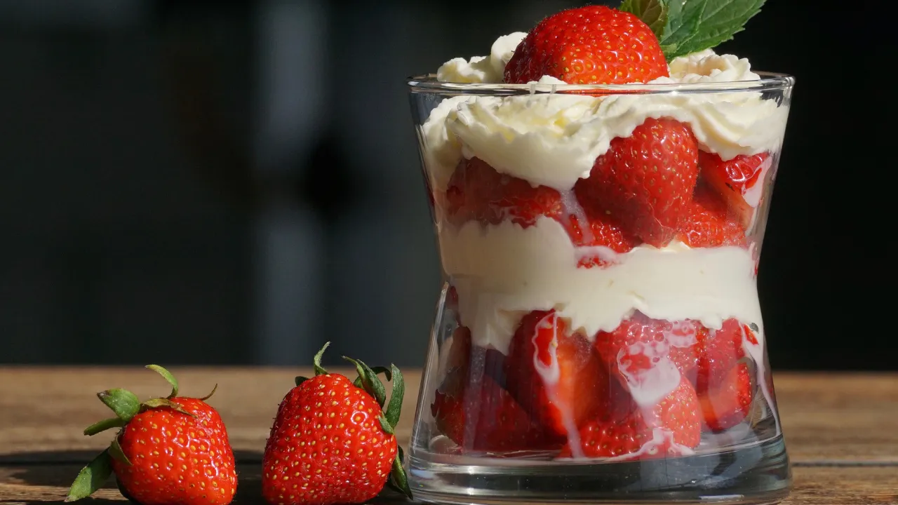 Sobremesa de morango fácil servida em copo transparente, com camadas de morangos frescos e creme branco, decorada com um morango inteiro e folha verde no topo. Dois morangos inteiros estão ao lado do copo sobre uma superfície de madeira.