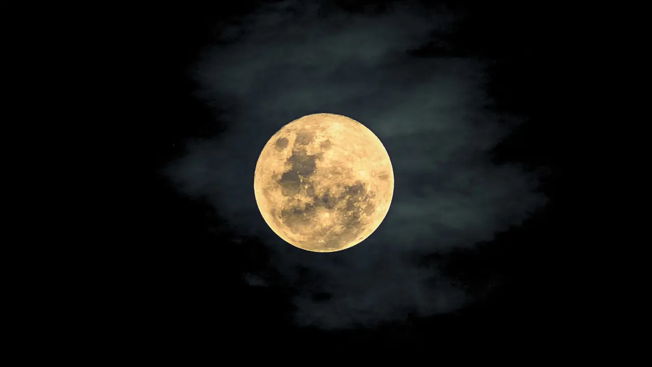 Imagem da lua de morango brilhando intensamente no céu noturno, parcialmente coberta por nuvens escuras, com um tom amarelado e texturas visíveis na superfície.